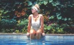 woman in bathing suit and sunglasses sits on pool ledge with her legs in the water